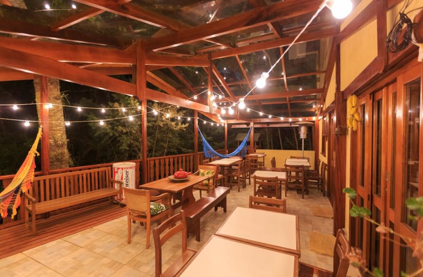 Wooden outdoor patio with string lights, dining tables, bench seating, hammock in back, warm night setting, tiled floor