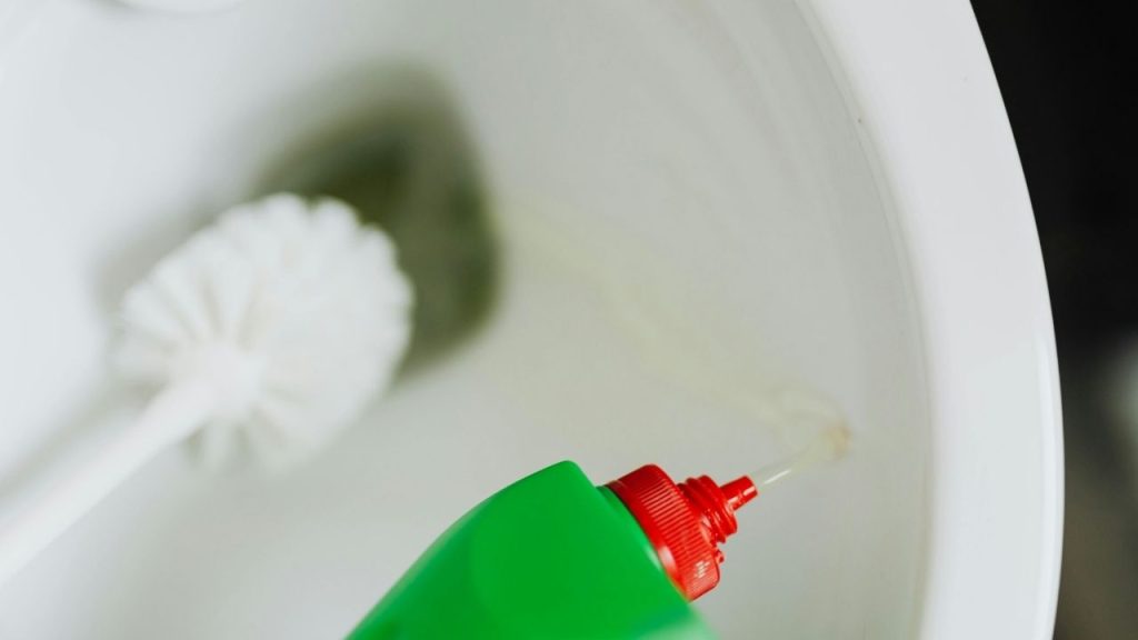 Green bottle with red nozzle, liquid cleaner being poured, toilet cleaning in progress disinfecting bathroom surfaces, household cleaning product, removing stains and germs, close-up of cleaning process
