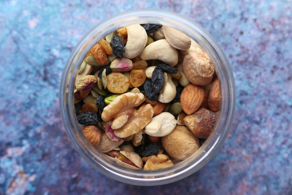 A top-down view of a glass jar filled with trail mix, including nuts, dried fruits, and seeds. The mix contains almonds, cashews, walnuts, pistachios, golden and black raisins, pumpkin seeds, and hazelnuts, against a textured blue background