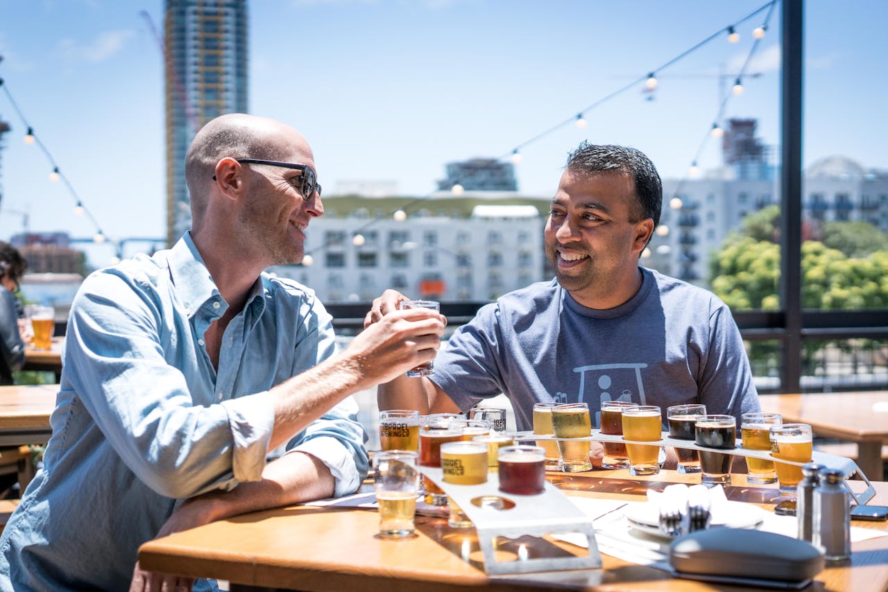 Two men sitting at an outdoor table, clinking glasses, smiling, wearing casual shirts, a flight of beers in front of them, city buildings in the background