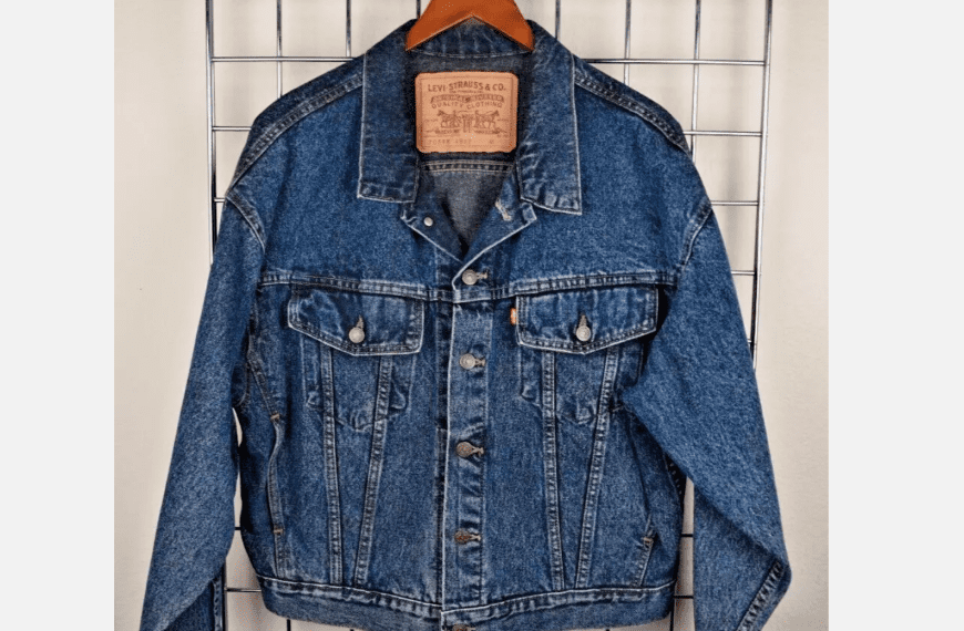 Blue Levi’s denim jacket hanging on a wooden hanger, displayed against a white grid wall, visible leather brand patch