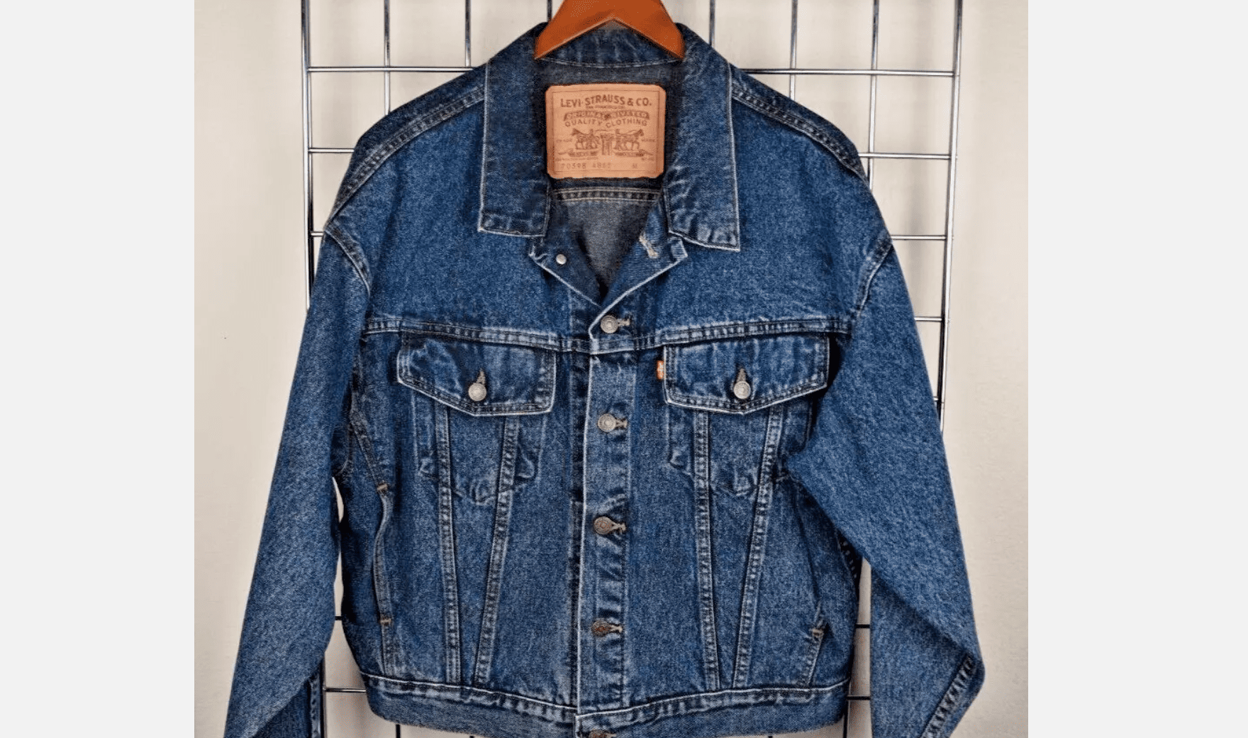 Blue Levi’s denim jacket hanging on a wooden hanger, displayed against a white grid wall, visible leather brand patch