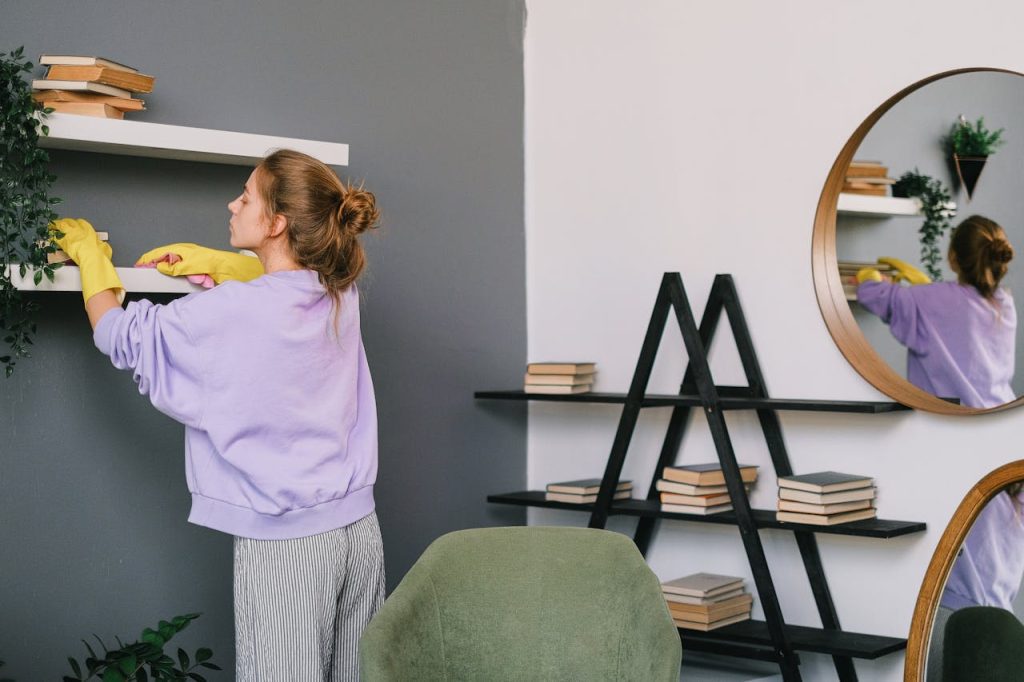 Woman cleaning a white shelf, wearing yellow gloves, adjusting or dusting books and decorations, modern interior with dark and light walls, round mirror reflecting the room