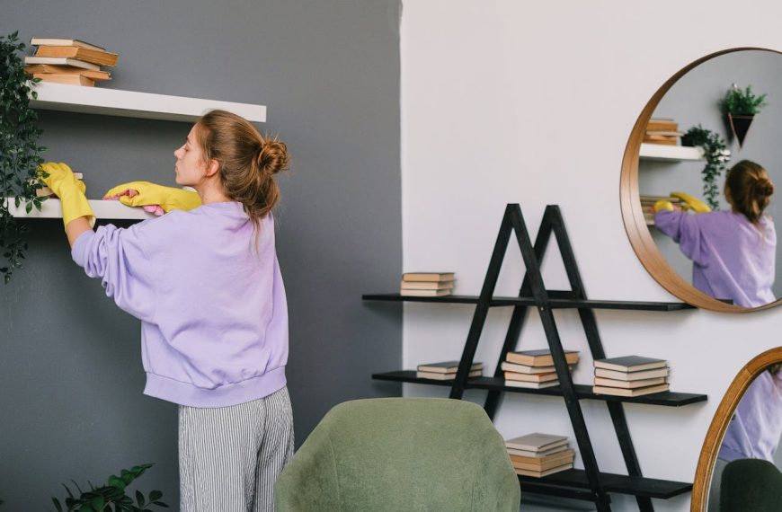Woman cleaning a white shelf, wearing yellow gloves, adjusting or dusting books and decorations, modern interior with dark and light walls, round mirror reflecting the room