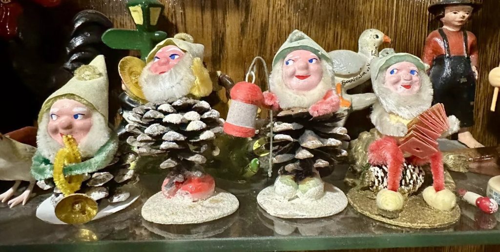 Vintage gnome or elf Christmas decorations made with pinecones, pipe cleaners, and glitter, displayed on a shelf with other retro figurines in the background