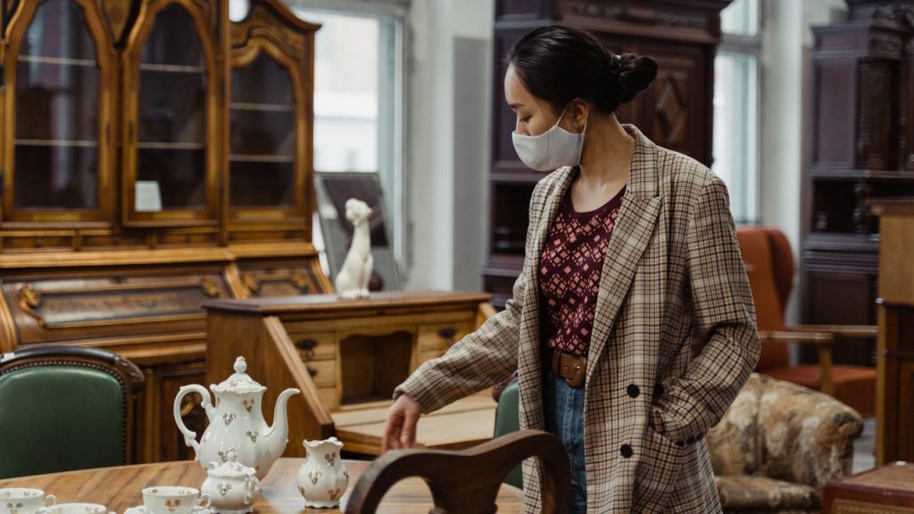 Woman in plaid coat wearing mask, examining porcelain tea set, antique furniture and cabinets in background
