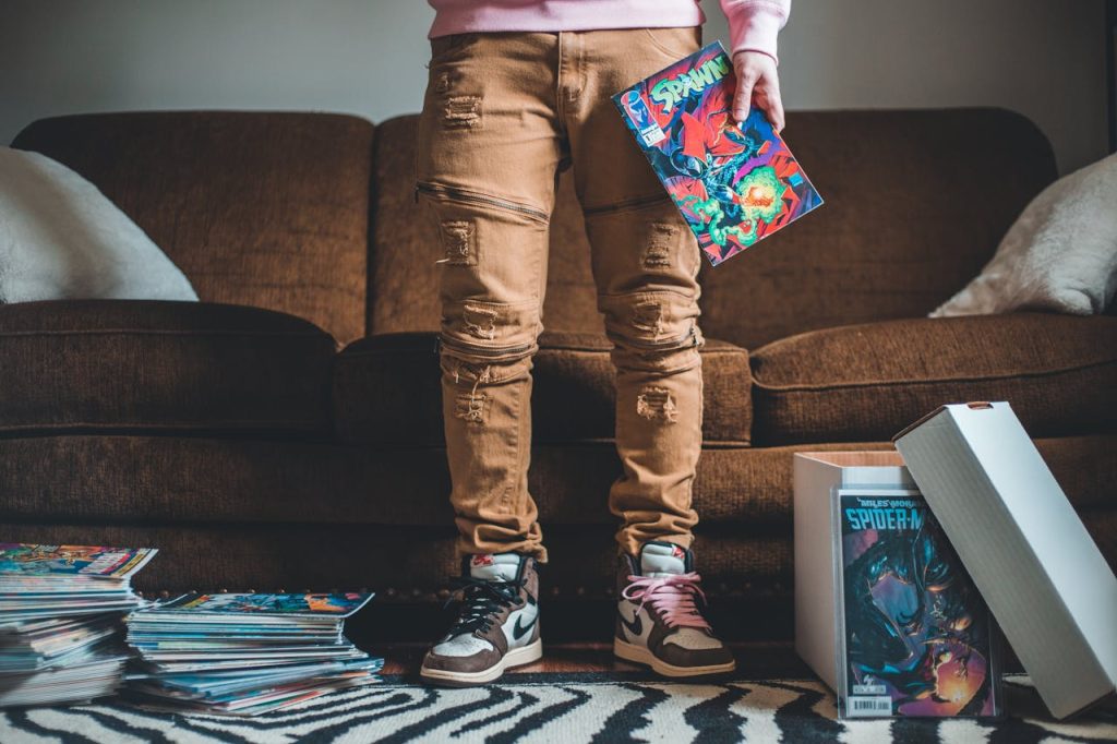 Person standing on zebra-print rug, holding Spawn comic, sneakers with mismatched laces, comic stacks and Spider-Man comic in white storage box, comic books