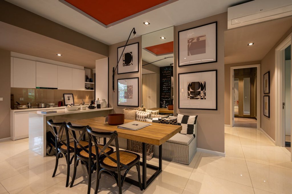 Modern open-plan apartment, wooden dining table with black chairs, kitchen in background, wall art, built-in bench seating, neutral tones with red ceiling accent