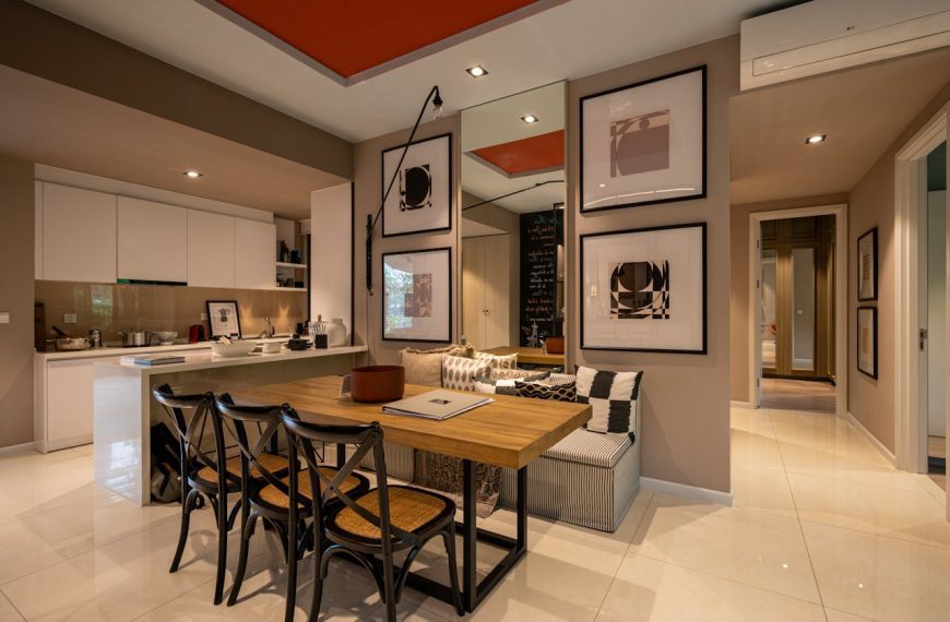 Modern open-plan apartment, wooden dining table with black chairs, kitchen in background, wall art, built-in bench seating, neutral tones with red ceiling accent
