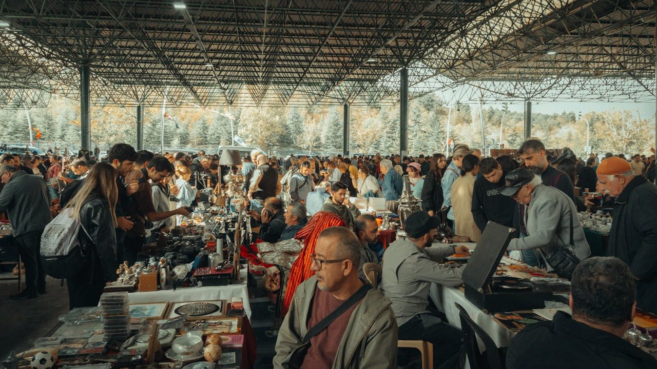 indoor flea market crowd, tables filled with vintage items, people browsing, high ceiling structure, daylight shining in