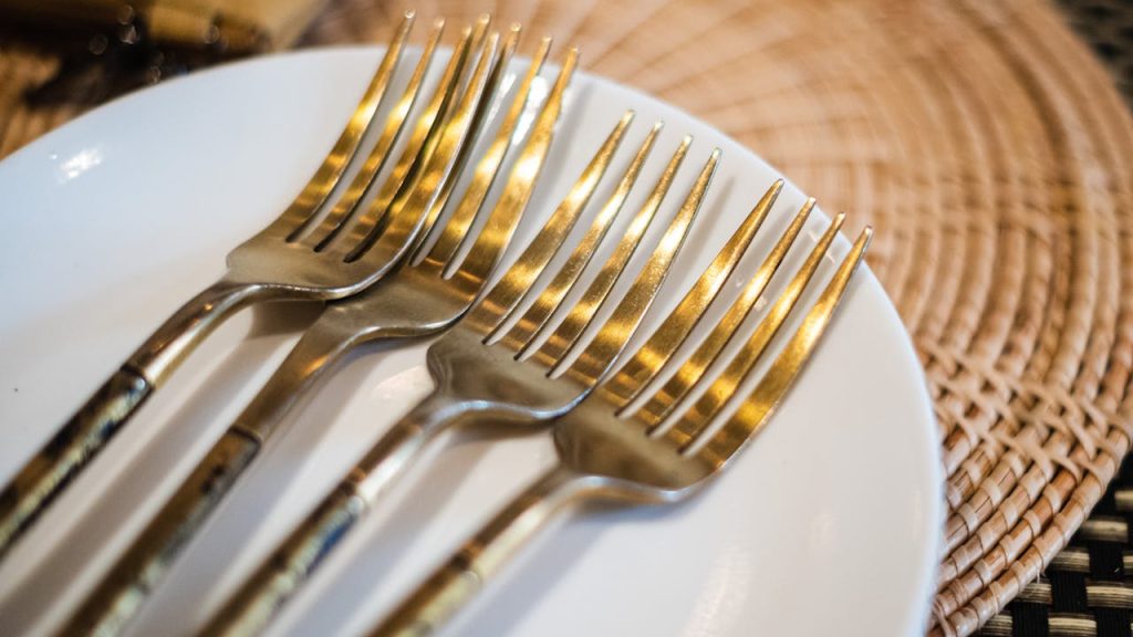 Four gold-toned forks neatly placed on a white plate, round woven placemat underneath, elegant table setting