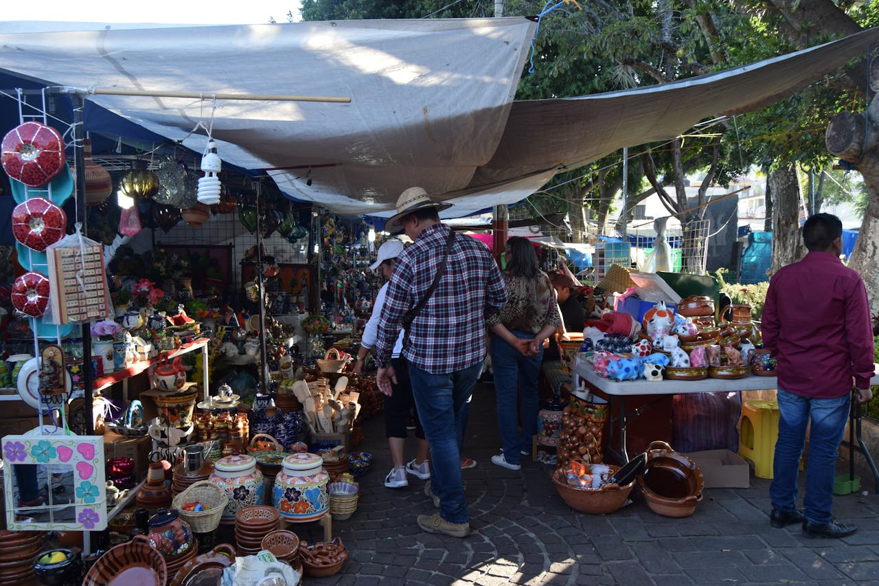 outdoor flea market, people browsing handmade pottery and crafts, colorful ceramics, shaded stalls, sunny day, trees in background