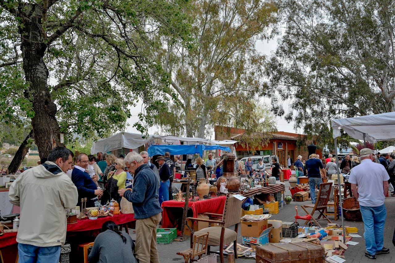 busy outdoor flea market, people browsing antique and vintage items, colorful stalls, shaded by large trees, casual daytime setting