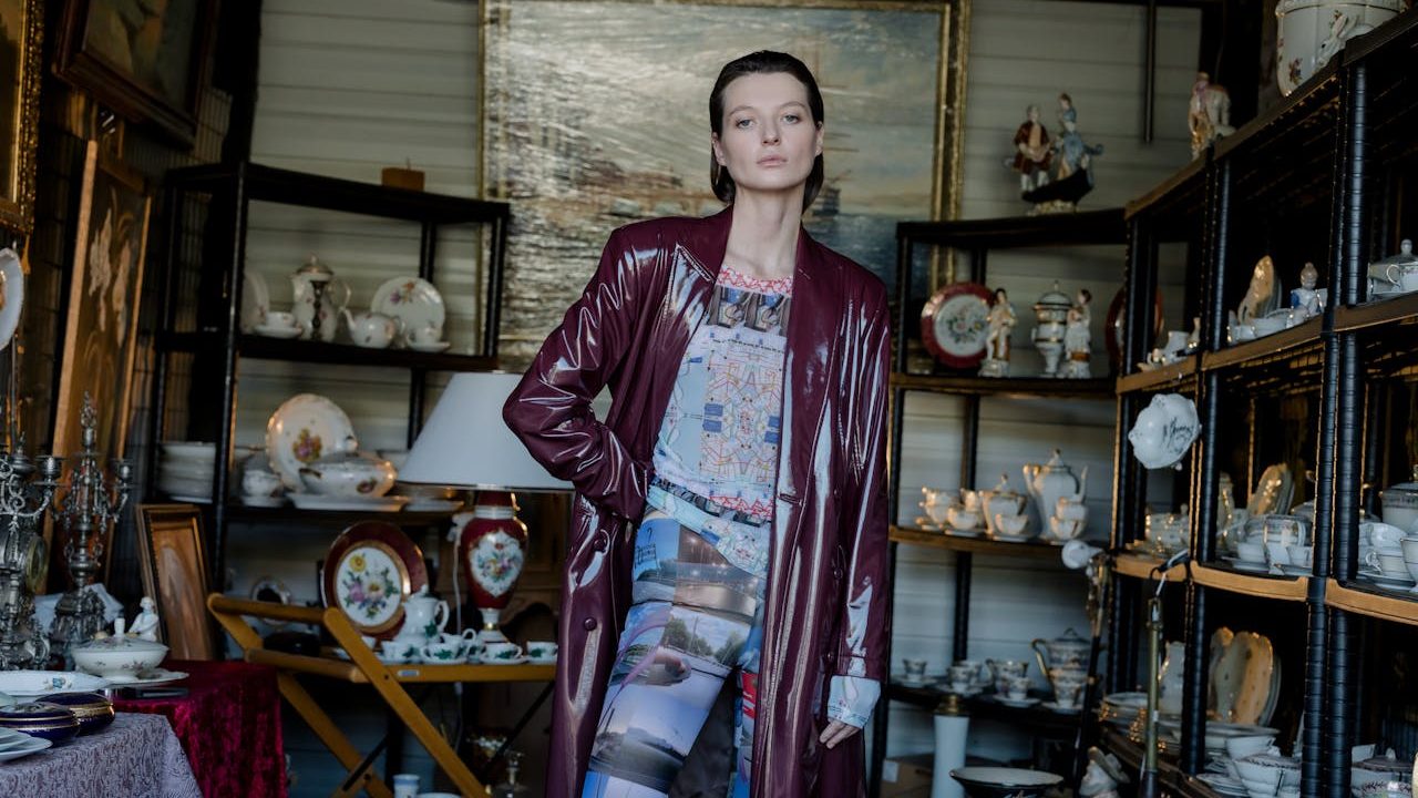A woman wearing a long burgundy leather coat and patterned pants stands confidently in a room filled with antique porcelain dishes, teacups, and decorative items on shelves behind her