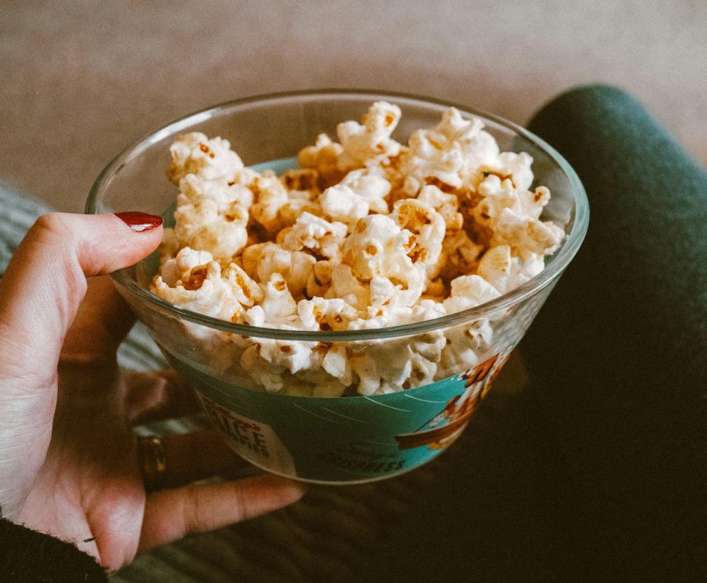 Hand holding a glass bowl filled with caramel popcorn, person sitting with legs crossed, cozy indoor setting