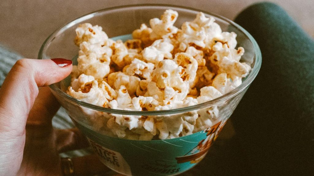 Hand holding a glass bowl filled with caramel popcorn, person sitting with legs crossed, cozy indoor setting