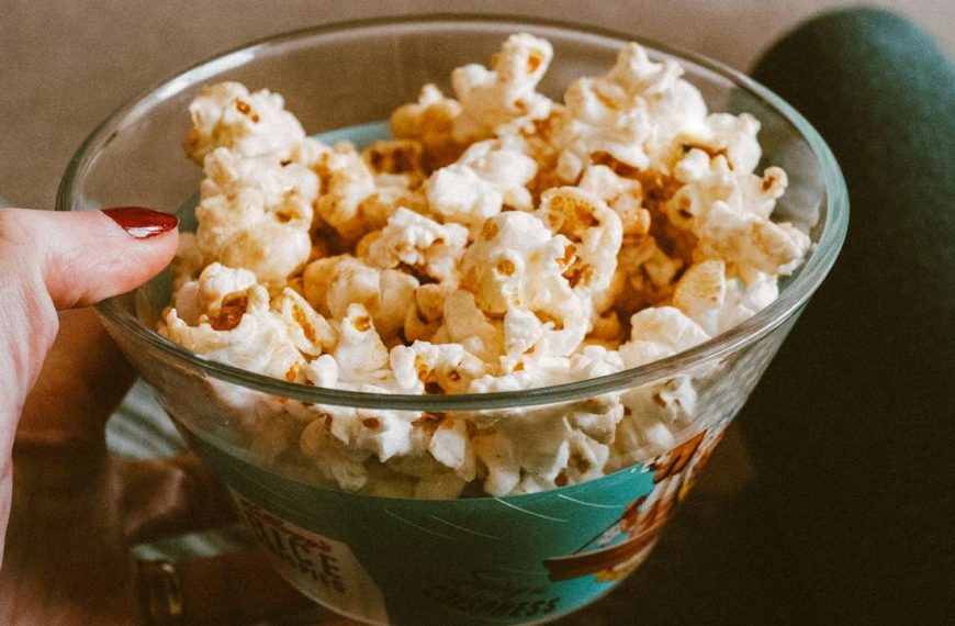 Hand holding a glass bowl filled with caramel popcorn, person sitting with legs crossed, cozy indoor setting