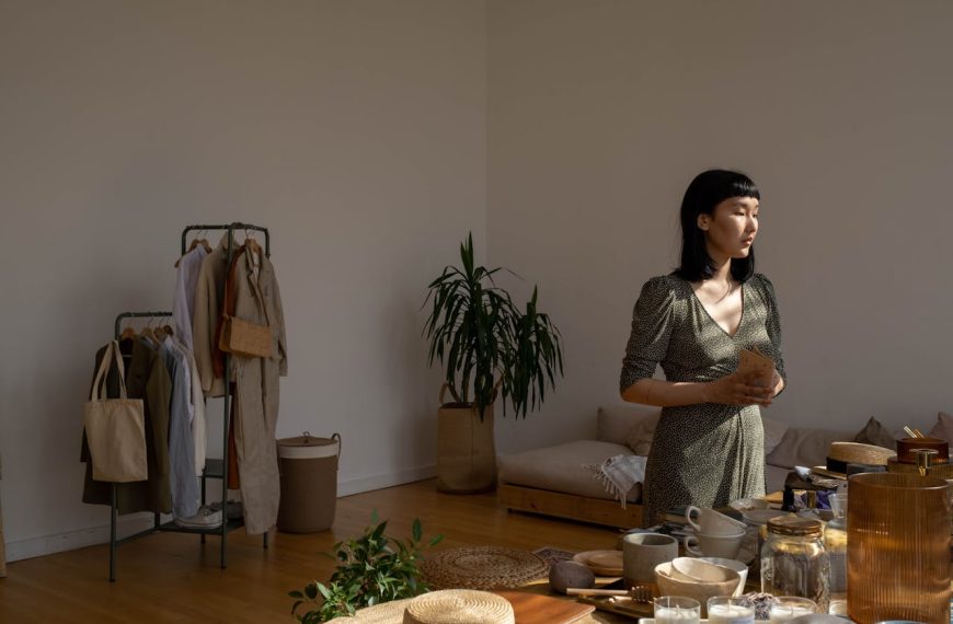 Woman standing in a sunlit, minimalist room holding a cup, with plants, clothing rack, and low furniture in the background