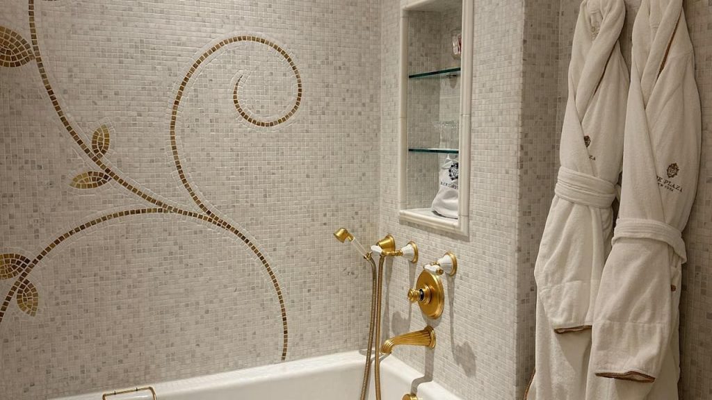 Luxury bathroom with marble mosaic walls featuring gold vine pattern, brass fixtures, built-in shelving, and white bathrobes
