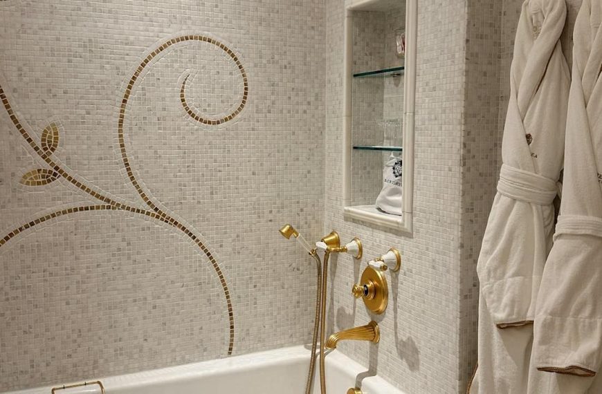 Luxury bathroom with marble mosaic walls featuring gold vine pattern, brass fixtures, built-in shelving, and white bathrobes