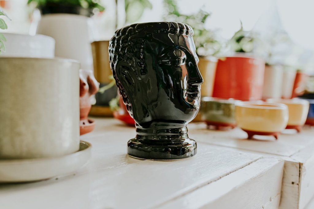 Black ceramic mug shaped like a head, surrounded by colorful cups and planters, displayed on a white shelf, natural light
