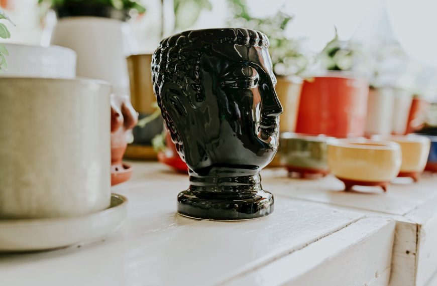 Black ceramic mug shaped like a head, surrounded by colorful cups and planters, displayed on a white shelf, natural light