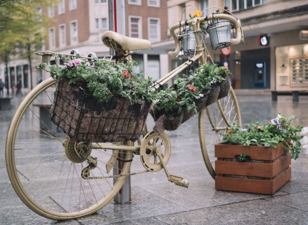 Vintage bicycle painted gold, filled with flower baskets, wooden crate with plants nearby, urban street setting, wet pavement