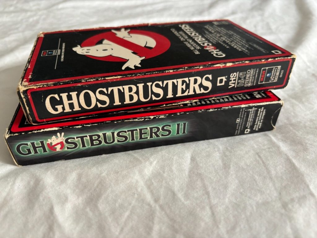 Ghostbusters and Ghostbusters II VHS tapes, stacked on white fabric, bold title fonts, classic 80s movie covers visible