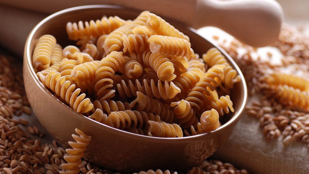 Brown ceramic bowl filled with spiral pasta and melted cheese, likely mac and cheese or a similar dish. The bowl sits on what appears to be a wooden surface. The image has a blue border
