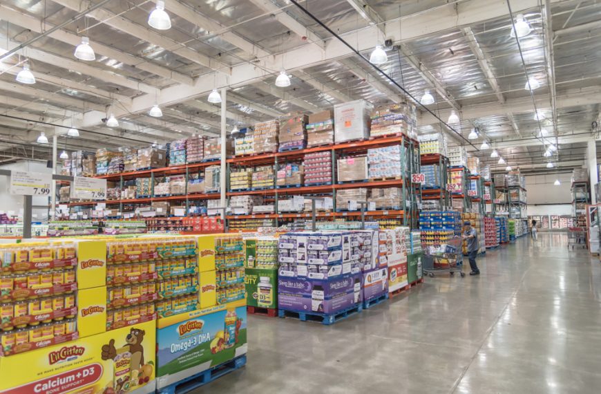 large warehouse store interior, tall industrial shelves stacked with bulk groceries, polished concrete floor, shopper walking down aisle