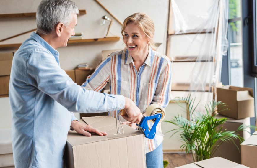 Smiling older couple taping cardboard box, light-filled room, indoor plants, casual clothing, home interior with shelves