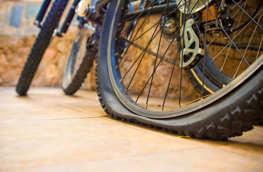 Close-up of a bicycle with a completely flat rear tire, resting on tiled floor, background showing other parked bikes and a stone wall