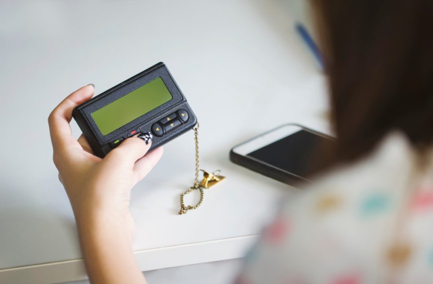 Person holding a black pager with a chain and charm, smartphone lying on a white desk nearby