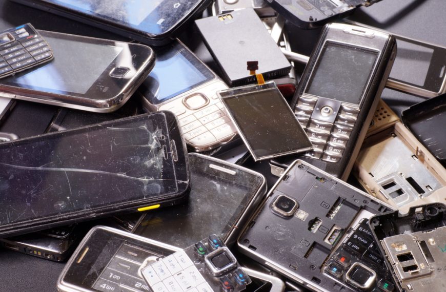 Pile of old, broken, and disassembled mobile phones with cracked screens, missing buttons, and exposed circuit boards