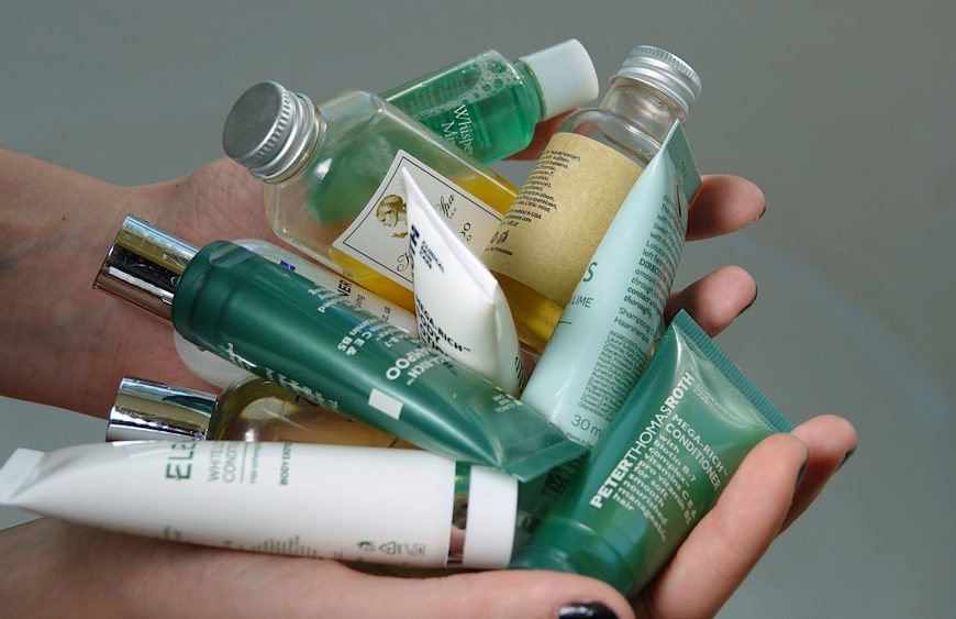 Hands holding assorted travel-size toiletry bottles and tubes, including shampoos, conditioners, and lotions