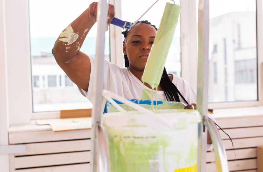 Woman loading paint roller with light green paint, standing by ladder, paint bucket in foreground, white room interior