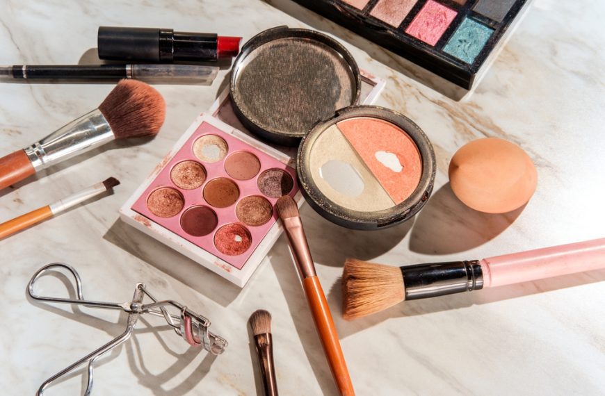 Various makeup products including eyeshadow palettes, blush, brushes, and lipstick scattered on a marble surface
