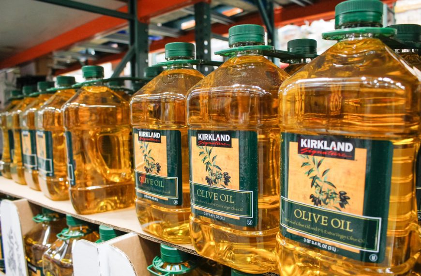 Large bottles of Kirkland Signature olive oil, green caps, arranged on a store shelf, bright lighting, wholesale warehouse setting, product labels visible, clear golden liquid inside