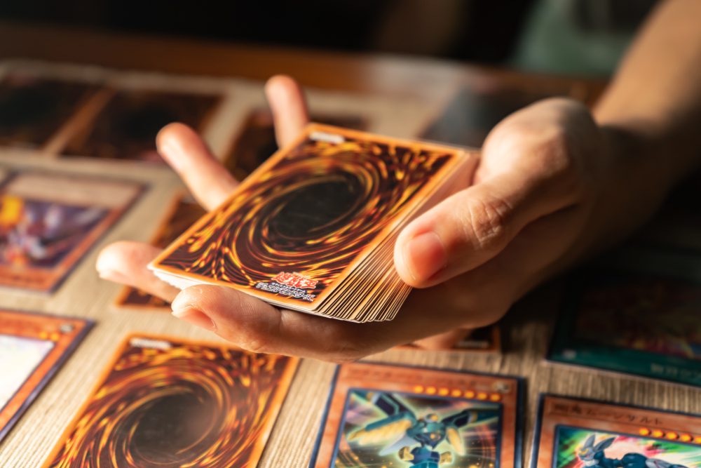 A hand holding a deck of Yu-Gi-Oh! trading cards above a table spread with face-up cards
