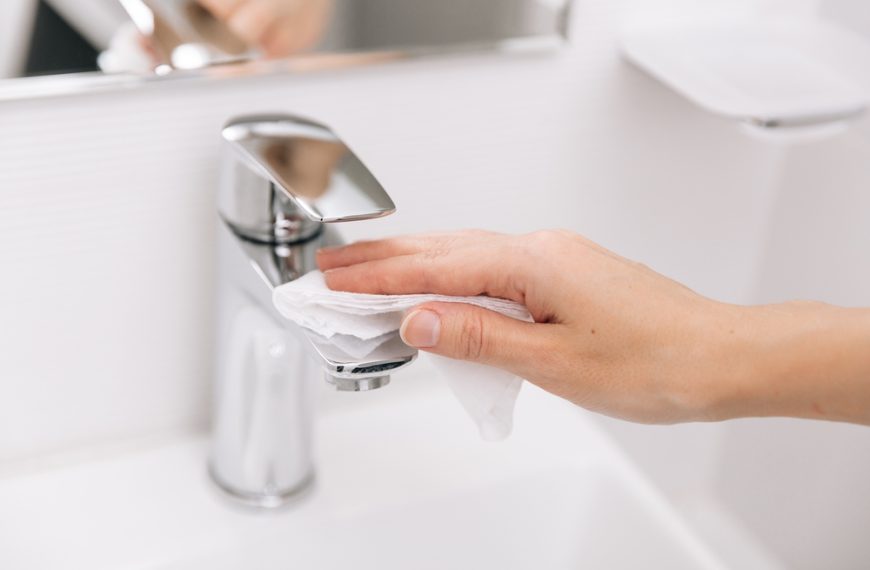 Hand wiping chrome faucet with a white cloth, modern sink and mirror in background, bathroom setting