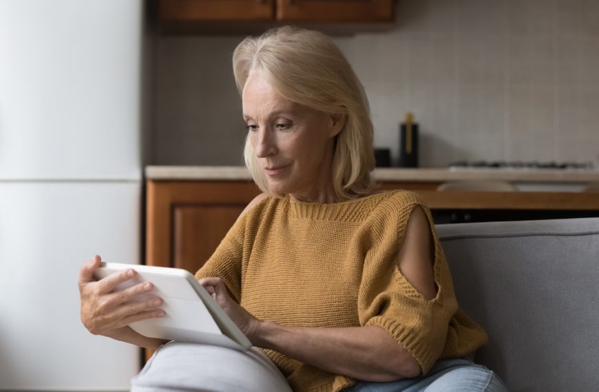 Older woman sitting in kitchen using a tablet, wearing mustard sweater, focused expression, casual setting