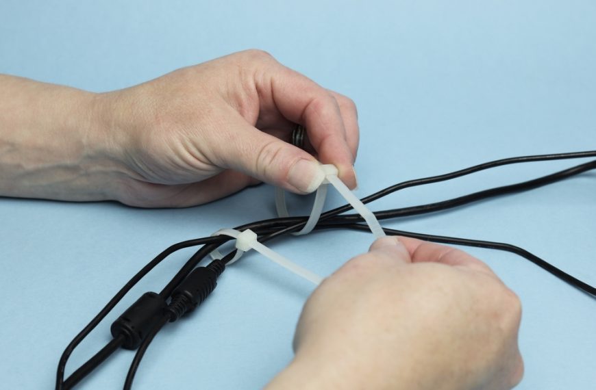 Hands tying plastic zip ties around multiple black cables to organize and manage wires on light blue background