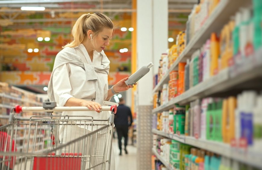 Woman shopping in grocery store, reading product label, pushing shopping cart, standing in cereal aisle