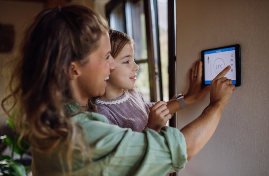 Smiling woman and young girl adjusting a smart thermostat on a wall, bright natural light from a nearby window