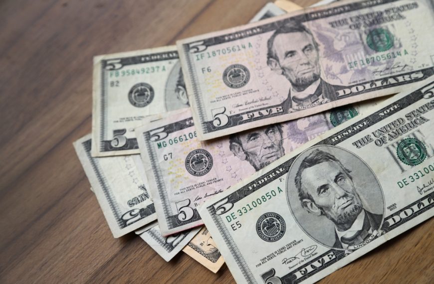 Scattered five-dollar bills on wooden surface, showing Abraham Lincoln portrait, slightly overlapping arrangement, U.S. currency