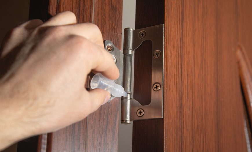 Hand applying lubricant to metal door hinge, wooden door frame, close-up view, home maintenance in progress
