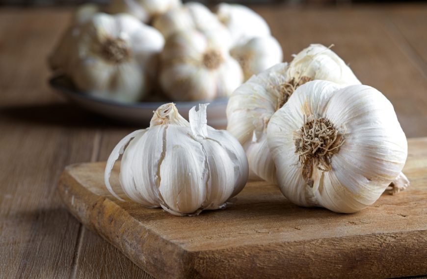 whole garlic bulbs on a wooden cutting board with more garlic in a bowl in the background