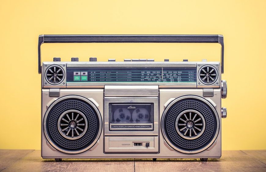 Vintage silver boombox radio with dual speakers, cassette deck, and control buttons, placed against a solid yellow background
