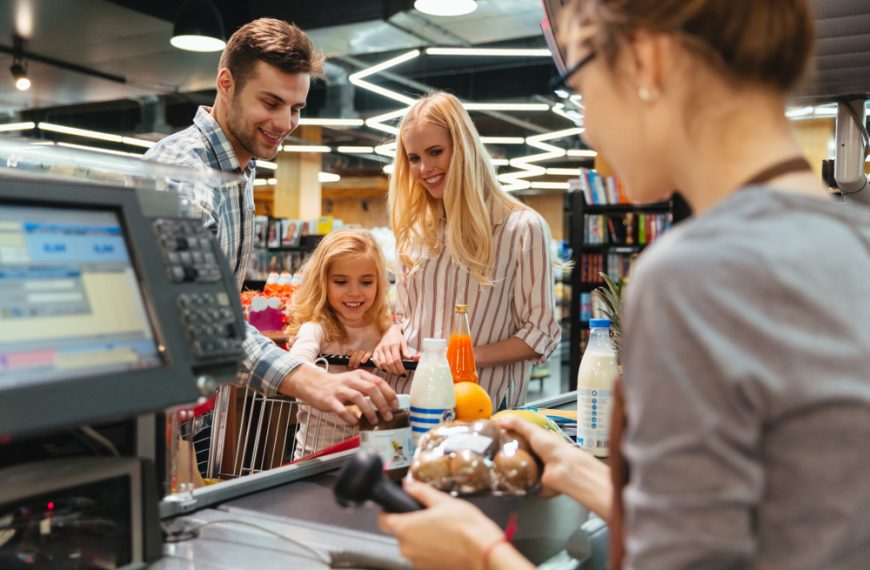 Smiling family at supermarket checkout counter, cashier scanning groceries, shopping cart filled with food items