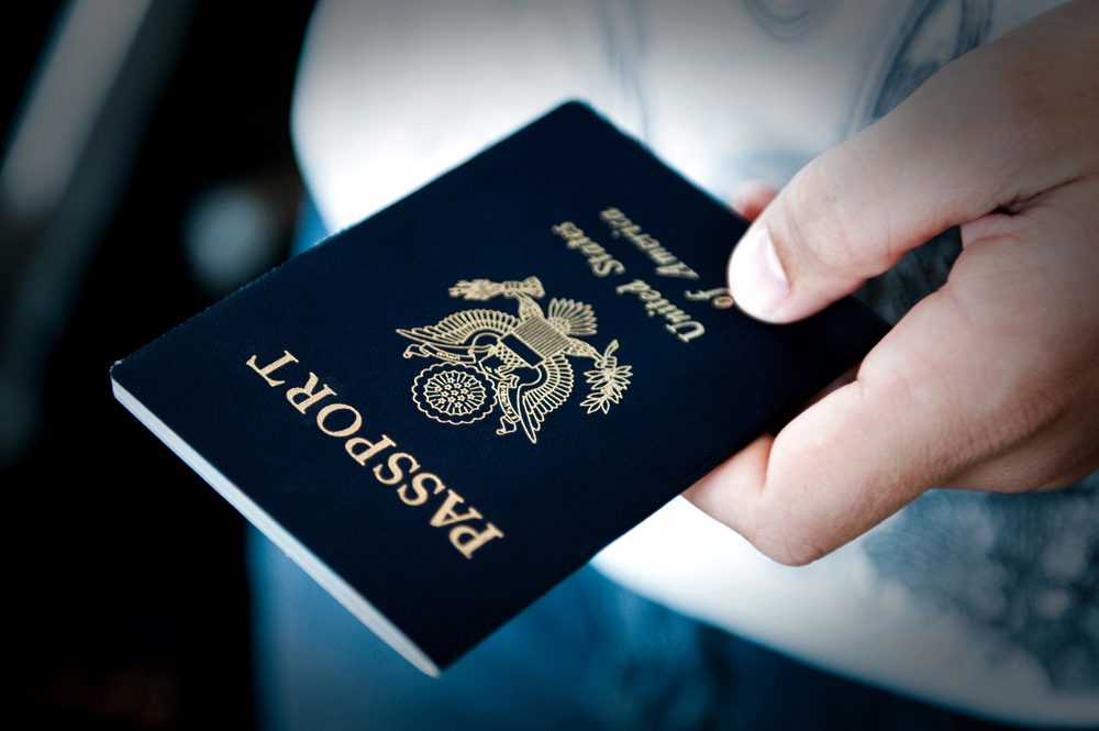 Hand holding a dark blue passport, gold UK coat of arms visible, text reads "PASSPORT" and "United Kingdom of Great Britain and Northern Ireland", person wearing light-colored shirt and jeans, close-up view, blurred background